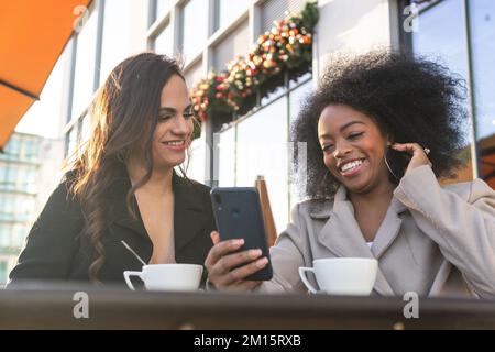 From below of laughing black businesswoman sharing mobile phone with diverse female partner while having coffee in downtown Stock Photo
