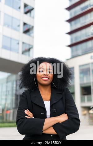 Confident African American female entrepreneur in smart casual clothes with curly hair crossing arms and looking at camera while standing near skyscra Stock Photo
