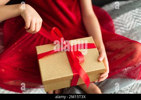 From above cropped unrecognizable girl in red dress sitting cross legged on bed and pulling red ribbon on golden box during holiday celebration at hom Stock Photo