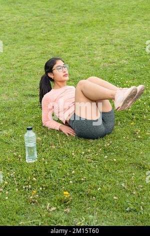 From above of young female in sports clothes and eyeglasses with plastic bottle of water lying on grass with legs raised while doing exercise during w Stock Photo