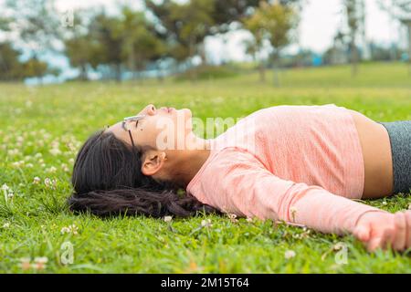 Side view of young fit female in pink long sleeved top and glasses with dark hair resting on green grass after intense training Stock Photo