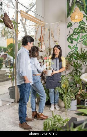 Full body of young couple choosing potted plant in floral shop while female florist in apron helping with selection Stock Photo