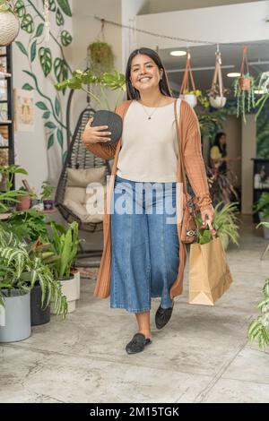 Full body of positive female customer in stylish outfit smiling and looking at camera while carrying potted plants in cozy flower shop Stock Photo