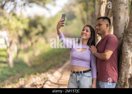 Happy young couple in casual clothes taking selfie with smartphone while standing near tree in park Stock Photo