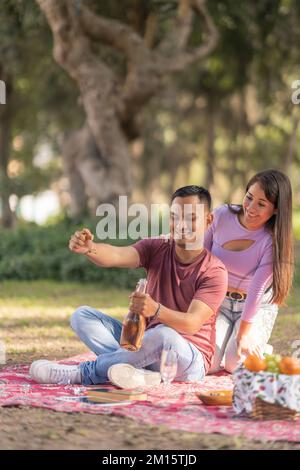 Young Hispanic woman looking at boyfriend opening bottle of wine while having picnic on blanket in park during weekend Stock Photo