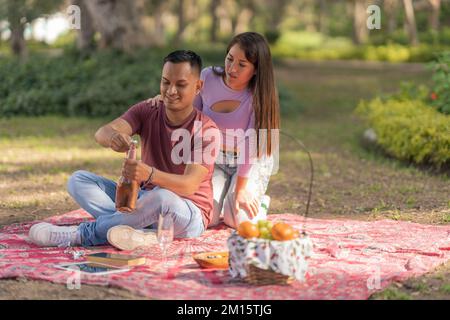 Young Hispanic woman looking at boyfriend opening bottle of wine while having picnic on blanket in park during weekend Stock Photo
