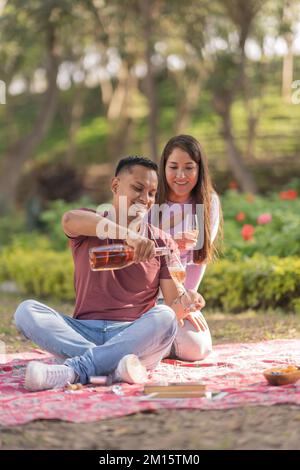 Young Hispanic woman looking at boyfriend opening bottle of wine while having picnic on blanket in park during weekend Stock Photo