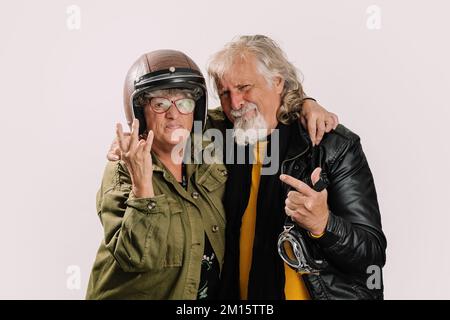 Cheerful senior woman in motorcycle helmet and khaki jacket showing three fingers gesture and hugging smiling aged male biker in leather jacket isolat Stock Photo