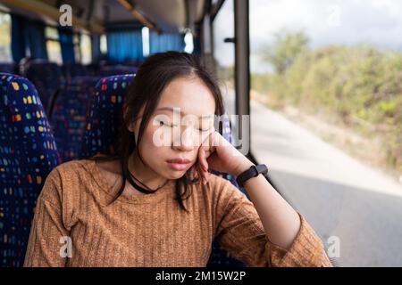 Sleeping young Asian female in brown sweater with smart watch sitting on comfortable passenger seat and leaning on arm while riding in Noto city Stock Photo