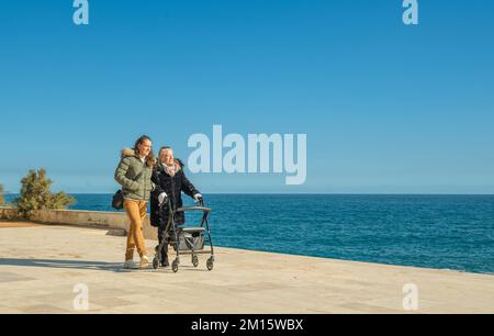 Full body of young woman and female pensioner with walkers in warm clothes walking on paved embankment near rippling seawater under cloudless blue sky Stock Photo