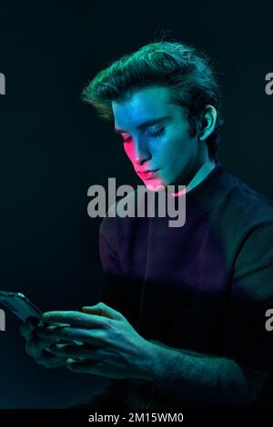Concentrated male in casual clothes browsing mobile phone while standing on black background with glowing blue and pink neon lights Stock Photo