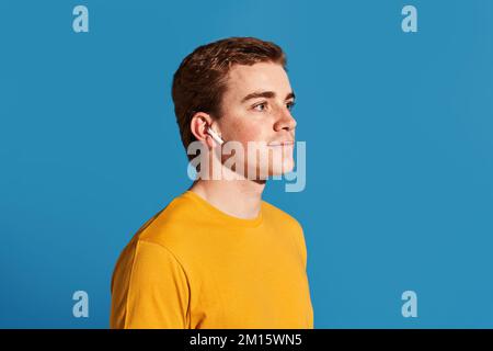 Side view of young redhead man in yellow t shirt touching true wireless earphone in ear and looking away while listening to music against blue backgro Stock Photo