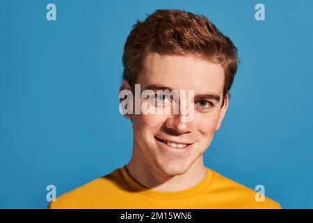 Happy redhead young male in yellow t shirt looking at camera against blue background Stock Photo