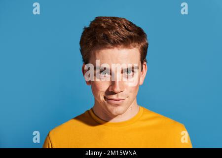 Redhead young male in yellow t shirt smirking and looking at camera against blue background Stock Photo