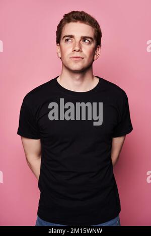 Thoughtful young male in black t shirt looking away standing against pink background Stock Photo