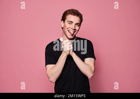 Happy young male in black t shirt clasping hands and showing tongue against pink background Stock Photo