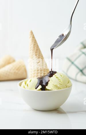 Unrecognizable person pouring chocolate sauce on scoops of delicious vanilla ice cream served in bowl and decorated with waffle cone on kitchen table Stock Photo