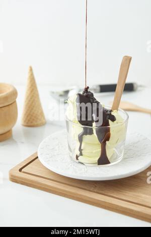 High angle of delicious chocolate syrup pouring on yummy homemade ice cream served in glass bowl on wooden cutting board near waffle cone in kitchen Stock Photo