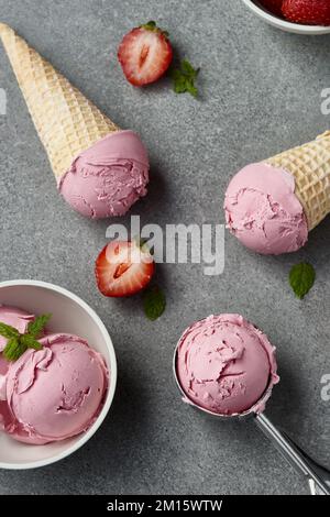 Top view of yummy berry ice cream in waffle cones placed on gray table near metal scoop bowl and strawberries in kitchen Stock Photo