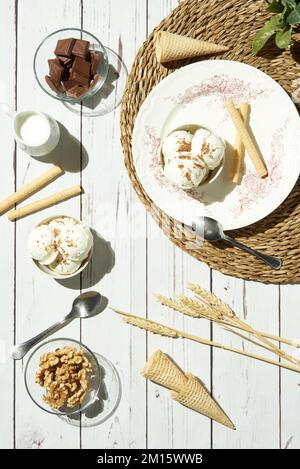 From above of delicious ice cream served in bowls with waffle rolls and cones and placed on white wooden table near pitcher with milk chocolate bars a Stock Photo