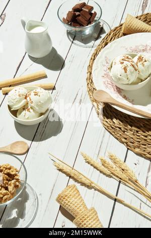 From above of delicious ice cream served in bowls with waffle rolls and cones and placed on white wooden table near pitcher with milk chocolate bars a Stock Photo