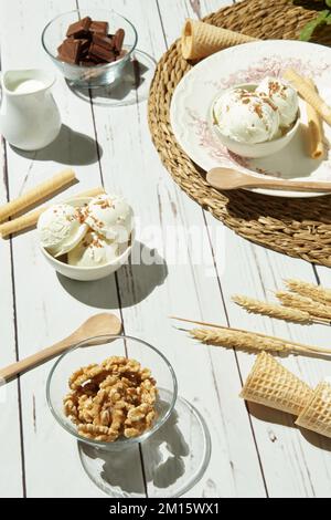 From above of delicious ice cream served in bowls with waffle rolls and cones and placed on white wooden table near pitcher with milk chocolate bars a Stock Photo