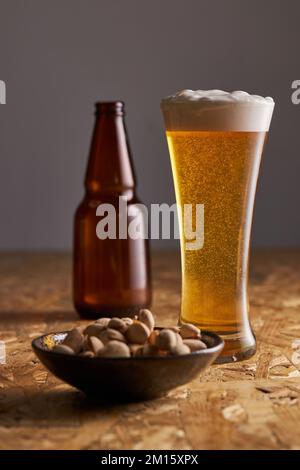Glass of fresh cold beer placed on wooden table near bottle and plate with pistachios Stock Photo