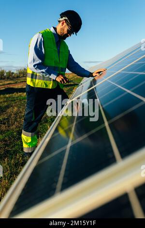 Side view of focused technician in uniform working with solar panels in grassy countryside under cloudless blue sky in sunlight Stock Photo