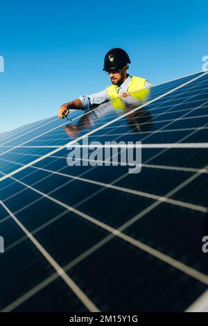 Low angle of bearded worker in protective helmet with screwdriver installing photovoltaic cells against blue sky Stock Photo