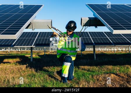 Worker in uniform and protective helmet connecting wires while installing photovoltaic cells in field on sunny day Stock Photo
