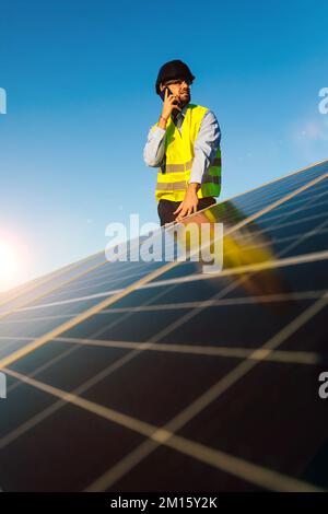 Worker in helmet and uniform looking away while standing near photovoltaic cells against cloudless blue sky and speaking on cellphone Stock Photo