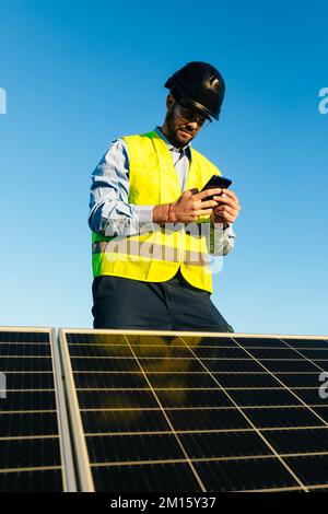 Low angle of positive male engineer in protective helmet and bright waistcoat standing near solar batteries and browsing cellphone on sunny day Stock Photo
