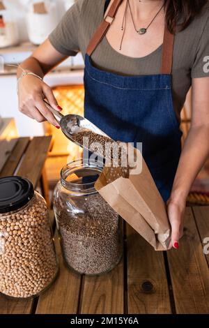 Crop unrecognizable female in apron standing at counter with scoop while pouring brown seeds into paper bag Stock Photo