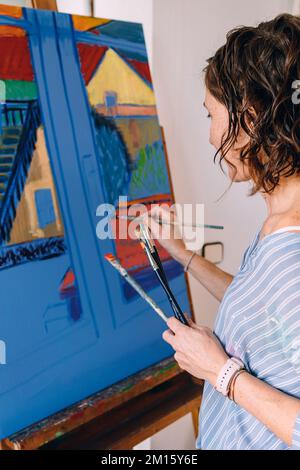Side view of young female in striped shirt standing near easel and painting on canvas with brushes Stock Photo