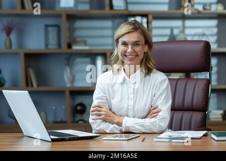 Successful businesswoman boss in shirt sitting at table with crossed arms smiling and looking at camera, lawyer working in modern office using laptop at work. Stock Photo