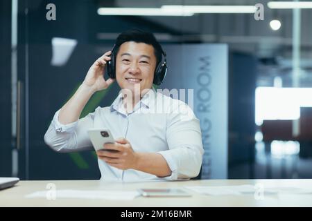 Portrait of a young handsome Asian man in the office. He is sitting at his desk, wearing headphones, holding a phone. He looks at the camera, smiles. Stock Photo
