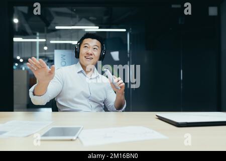 Joyful young Asian businessman listening to music in headphones at workplace. Holds the phone in his hands, sings, sits at the table and dances. Stock Photo