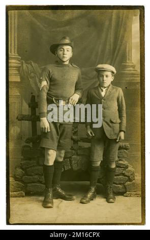 Original early 1920's postcard of two working class brothers - the eldest, a teenager, is dressed in  the  uniform  of the Royal Marines Volunteer Cadet Corps (formerly Royal Marines Artillery Cadet Corps until the 1910's). The 3 chevrons and a crown on his sleeve denote he was rank of Cadet Colour Sergeant. He is standing next to his younger brother. Both lads wear short trousers and boots, the youngest a jacket and flat cap. In the early 1920's short trousers and long socks became normal for boys and they wore them up until the age of 13 and the fashion continued up until the 1960's.  U.K. Stock Photo