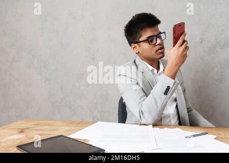 Young serious indian businessman using cellphone while working with paper documents in office Stock Photo