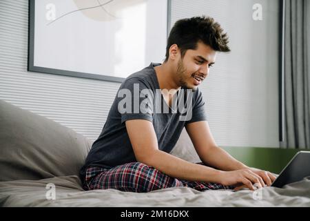 Young indian man with laptop sitting on bed and typing in cozy room at home Stock Photo
