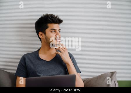 Young indian thoughtful man touching chin with his hand looking rightward sitting with laptop at home Stock Photo