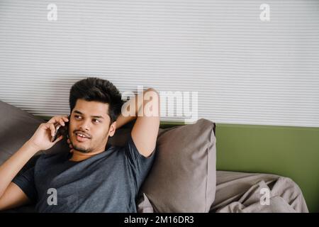 Young indian man liying on bed with hand behind his head and talking phone Stock Photo