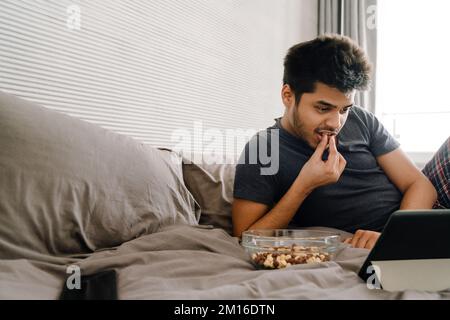 Young handsome indian man lying on bed with tablet eating snack and watching serial in cozy bedroom at home Stock Photo