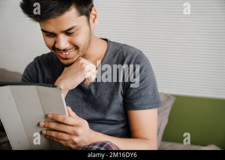 Young indian smiling man touching his chin sitting on bed with tablet at home Stock Photo