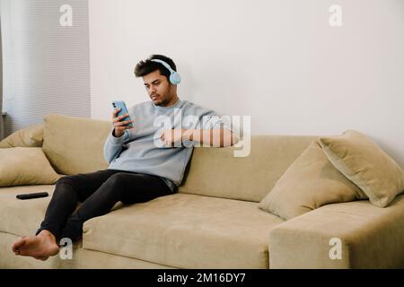 Young indian man in headphones with phone sitting on couch at home Stock Photo