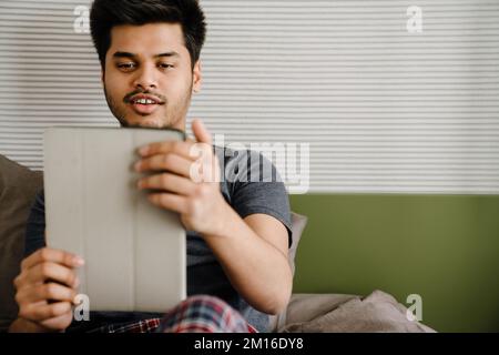 Young handsome smiling indian man holding tablet looking on it while sitting at home Stock Photo