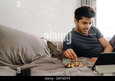 Young handsome smiling indian man lying on bed with tablet eating snack and watching serial in cozy bedroom at home Stock Photo