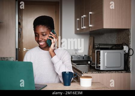 Cheerful black woman speaking on cellphone while using netbook in kitchen Stock Photo