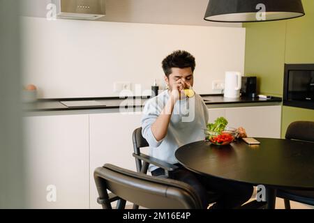 Young indian man drinking orange juice while having lunch with bowl of fresh salad Stock Photo