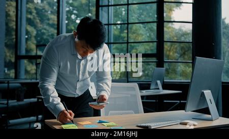Senior asian ethnic man entrepreneur businessman stand at table in modern office use mobile phone write tasks on sticky notes organize paperwork Stock Photo